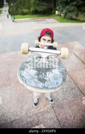 Ein junger langhaariger Skater in T-Shirt und durchgehender Cap steht mit einem Skateboard vor dem Hintergrund der Stadt Stockfoto