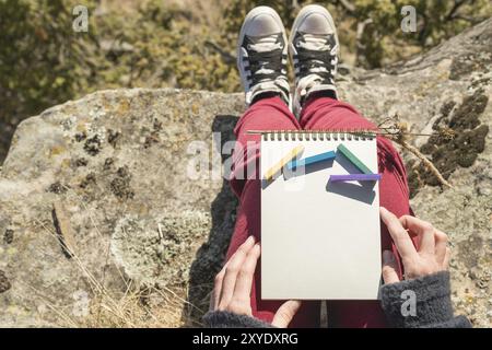 Blick von der ersten Person auf dem weiblichen Schoß ist ein Notizbuch mit Kreiden zum Zeichnen von Pastellfarben. Weibliche Hände, die ein Notizbuch in der Natur halten. Ein Mädchen sitzt Stockfoto