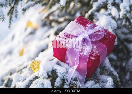 Rotes Neujahrsgeschenk mit weißem Band neben Weihnachtsspielzeug auf den Zweigen eines schneebedeckten Weihnachtsbaums im Winterwald. Der Kontra Stockfoto