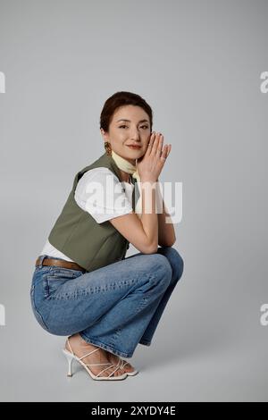 Eine stilvolle asiatische Frau mit grüner Weste und Halstuch posiert in der Hocke vor grauem Hintergrund. Stockfoto