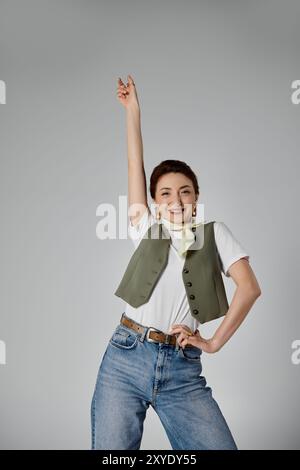 Eine stilvolle asiatische Frau mit Weste und Halstuch posiert vor grauem Hintergrund und lächelt selbstbewusst. Stockfoto
