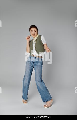 Eine stilvolle asiatische Frau in grüner Weste und weißem Halstuch posiert vor grauem Hintergrund und trägt blaue Jeans und weiße Sandalen. Stockfoto