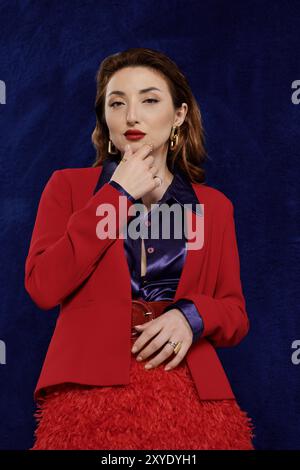Eine attraktive asiatische Frau in einem leuchtend roten Anzug und einem Seidenhemd posiert selbstbewusst vor einem dunkelblauen Hintergrund. Stockfoto