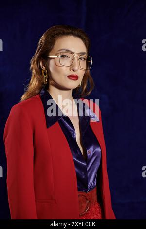 Eine stilvolle asiatische Frau in rotem Blazer und Seidenhemd posiert selbstbewusst vor dunklem Hintergrund. Stockfoto