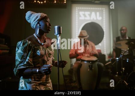 Junge Schwarze Frau in stilvollem Kleid mit Kopfbedeckung, die während des Auftritts mit einer Jazzband im Mikrofon singt Stockfoto