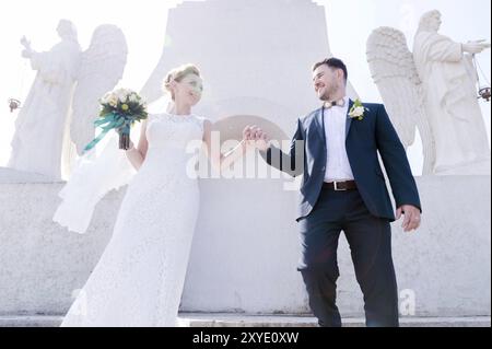 Porträt eines wunderbaren Paares, das an einem Hochzeitstag mit einem Blumenstrauß in der Hand vor dem Hintergrund eines orthodoxen christlichen Denkmals mit Engeln verbrachte. Stockfoto