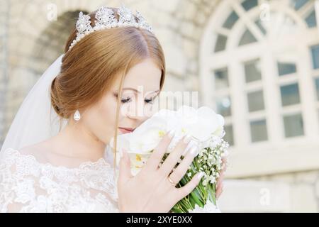 Lächelnde junge Braut in weißem Kleid schnüffelt an ihrem Hochzeitsstrauß, der sorgfältig von den Händen gehalten wird. Das Konzept der Floristen bei Hochzeiten Stockfoto