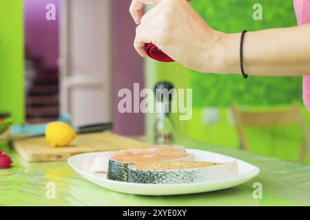 Frauenhände besprühen aus der Handmühle mit Gewürzen zwei rohe Steak vom Lachs, die auf einem weißen Teller in einer Küche in Grüntönen liegen. Das Konzept o Stockfoto