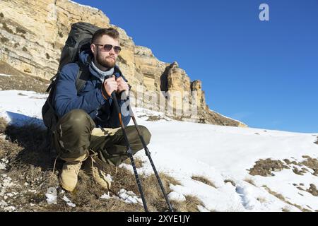 Porträt eines Hipster-Reisenden mit Bart in Sonnenbrille sitzt auf der Natur. Ein Mann wandert in den Bergen mit einem Rucksack und skandinavischem Walking sti Stockfoto