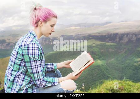 Ein Reisender sitzt in den Bergen auf dem Gras und liest ein Buch über den Hintergrund epischer Berge. Der Begriff des Lesens während der Ruhe und im Urlaub Stockfoto