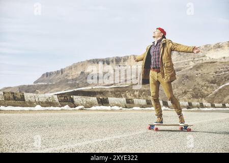 Stylischer Happy Young man in einer Mütze und Hose Jogginghose, die auf einem Longboard eine Bergstraße hinunterrollt und das Leben genießt Stockfoto