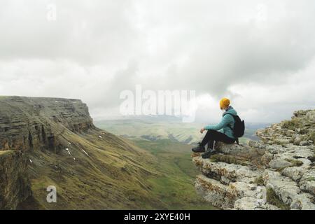 Junge Frau mit Rucksack, die pensiver am Rande eines Felsens sitzt und mit Wolken in den Himmel blickt Stockfoto