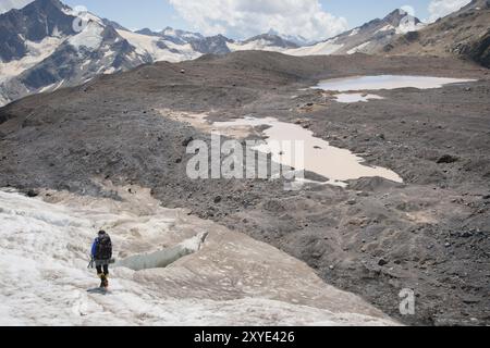 Ein Bergsteiger mit Rucksack läuft in Steigeisen entlang eines staubigen Gletschers mit Gehwegen in den Händen zwischen Rissen im Berg Stockfoto