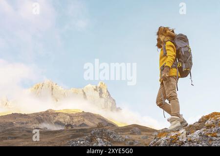 Das Backpacker-Mädchen in Sonnenbrille und einem großen Nordpelzhut mit einem Rucksack auf dem Rücken steht auf einem Felsen und blickt auf die Klippen, die sich darin verstecken Stockfoto