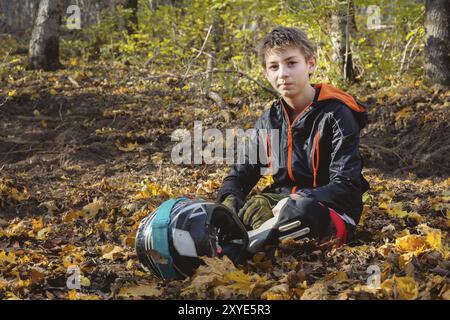 Porträt eines jungen Reiters, der im Wald auf einem gelben Laub sitzt und einen Vollhelm hält. Das Konzept des Outdoor-Sports für Kinder Stockfoto