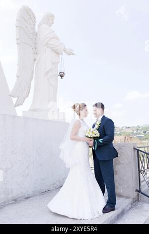 Porträt eines wunderbaren Paares, das an einem Hochzeitstag mit einem Blumenstrauß in der Hand vor dem Hintergrund eines orthodoxen christlichen Denkmals mit Engeln verbrachte. Stockfoto