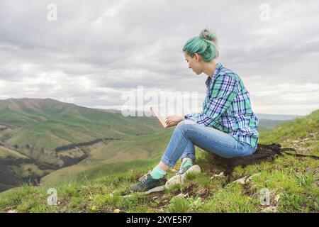 Porträt eines jungen Hipster-Mädchens mit mehrfarbigem Haar, das in den Bergen auf der Natur sitzt und ein Buch liest. Der Begriff des Lesens in der Natur. Bildungswesen Stockfoto