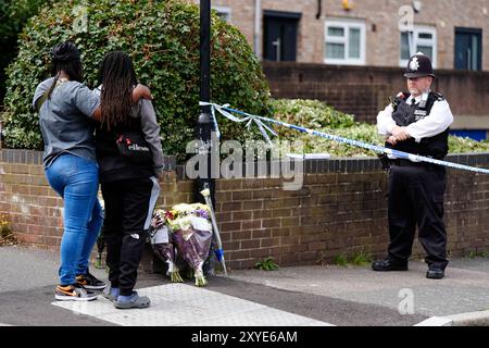 Ein Polizist steht neben einem Polizeikordel, während Mitglieder der Öffentlichkeit Blumenrabatte in der Overbury Street, nahe dem Tatort in der Rushmore Road, Clapton, im Osten Londons, ablegen, nachdem ein Mann in seinen 30ern erstochen wurde. Zwei Männer wurden verhaftet. Bilddatum: Donnerstag, 29. August 2024. Stockfoto