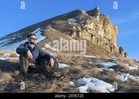 Porträt des ruhigen Hipster-Reisenden mit Bart in der Sonnenbrille sitzt auf der Natur. Ein Mann wandert in den Bergen mit Rucksack und skandinavischem Wa Stockfoto