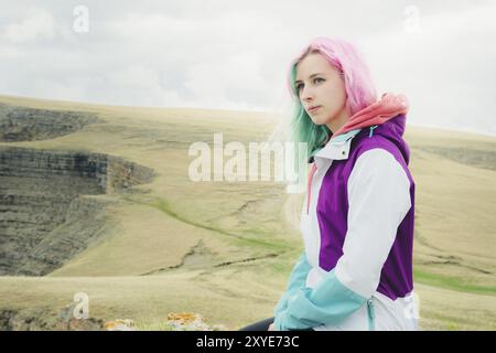 Ein Mädchen-Reisende mit bunten Haaren sitzt am Rande einer Klippe und blickt auf den Horizont auf einem Hintergrund eines felsigen Plateau Stockfoto