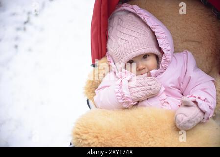 Cute Baby im Kinderwagen auf frostigen Wintertag. Baby tragen wollene warme Mütze und Handschuhe und Lügen in Lama Fell überzogen rot Kinderwagen. 1 Jahr altes Baby anhand von quantitativen Simulatio Stockfoto