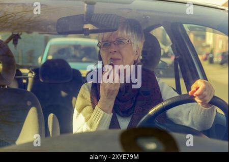 Die ältere Dame sitzt in ihrem Auto, schaut in den Rückspiegel und rührt ihren Lippenstift an Stockfoto