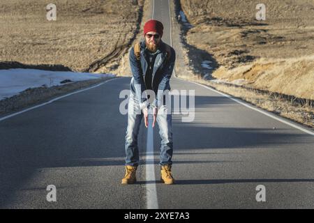 Ein stylischer bärtiger Hipster in Sonnenbrille mit einem Vintage-Rucksack steht an einem sonnigen Tag auf einem Landstraßenasphalt. Das Konzept des Anhalter- und Wanderns Stockfoto