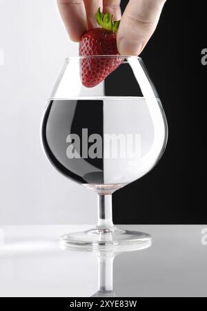 Hand mit einer roten Erdbeere über einem Glas klarer Flüssigkeit in zwei Teile mit einem künstlerischen Konzept des Brechungseffekts Stockfoto