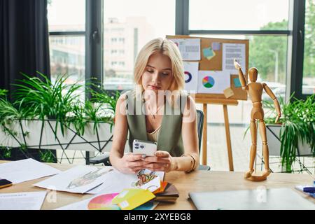 Eine blonde Frau in einem grünen ärmellosen Hemd sitzt an einem Schreibtisch in einem Büro, überprüft ihr Handy und sieht sich Designs auf dem Tisch an. Stockfoto
