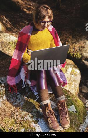 Ein getöntes Porträt eines lächelnden Freelancer-Hipster-Mädchens mit einer Brille in einer Decke und einem Laptop, der kniend auf einem Felsen in einem Nadelholz sitzt Stockfoto