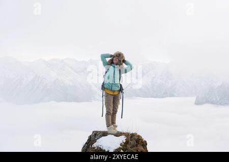 Ein fröhlich lächelndes Hipster-Mädchen reist in einer Daunenjacke mit Rucksack und in einem großen Pelzmütze steht auf einem Felsen vor dem Hintergrund eines verschwommenen M Stockfoto