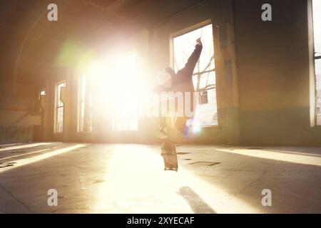 Ein junger Skater mit weißem Hut und schwarzem Sweatshirt macht einen Trick mit einem Skatesprung in einem verlassenen Gebäude im Hintergrund der untergehenden Sonne. Die Stockfoto