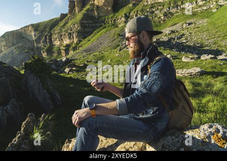 Nahaufnahme eines bärtigen Mannes in Jeanskleidung mit Sonnenbrille und einer Mütze mit Rucksack, der am Fuße der epischen Felsen sitzt und einen Kompass in der Hand hat. Stockfoto