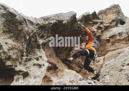 Hipster, Kletterer im Alter, in dem man einen wunderschönen Felsen ohne Versicherung und Helm hochklettert. Ein Bergsteiger in Hut und Daunenjacke mit Tasche für magnes Stockfoto