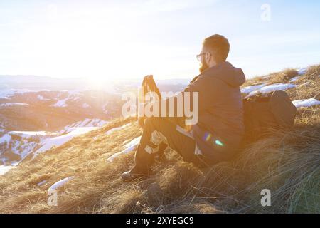 Hipster-Reisende mit Bart und Sonnenbrille sitzen im Winter auf einem gelben Gras auf einer Schieflage vor einem Hintergrund von schneebedeckten dales und einer bergigen CA Stockfoto