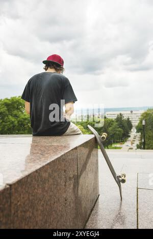 Der langhaarige Skater in Kappe und T-Shirt sitzt mit dem Rücken zur Kamera und denkt neben dem Skateboard vor dem Hintergrund der Landschaft Stockfoto