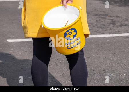 Freiwillige in gelbem Hemd mit Spendeneimer für Marie Curie Krebs-Wohltätigkeitsorganisation. Ballycastle, Großbritannien - 24. August 2024. Stockfoto