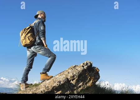 Stilvoller bärtiger Reisender in Sonnenbrille und einer Mütze mit Rucksack in Denim-Anzug und gelben Schuhen steht auf einem großen Stein vor dem Hintergrund Stockfoto