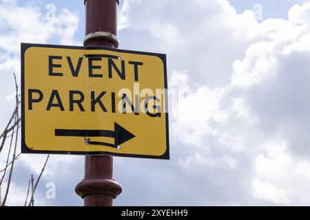 Gelbes Parkschild für Veranstaltungen mit schwarzer Schrift und Pfeil, der auf den ausgewiesenen Parkplatz an der Stange vor wolkenbewölktem, hellem Himmel zeigt Stockfoto