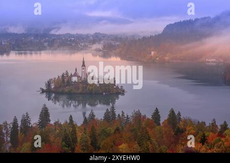 Morgen Antenne sonnenaufgang Blick auf den Bleder See mit der Kirche der Himmelfahrt der Maria, Slowenien im Nebel und Bäume im Herbst Hintergrund Stockfoto