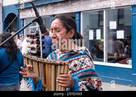 Mann mit langen Haaren und farbenfroher gemusterter Oberseite, der Pfannenpfeifen hält, traditionelle südamerikanische Flötenmusik. Ballycastle, Großbritannien - 26. August 2024. Stockfoto