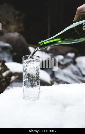 Mineralwasser wird aus einer glasgrünen Flasche in ein klares Glas-Becherglas gegossen. Ein Glas steht im Schnee. Vor dem Hintergrund eines Winters Stockfoto