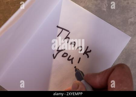 Eine Person schreibt eine Dankesnotiz auf ein Blatt Papier. Die Notiz befindet sich auf einem Holztisch und die Person schreibt sie mit einem Marker. Szene ist positiv Stockfoto