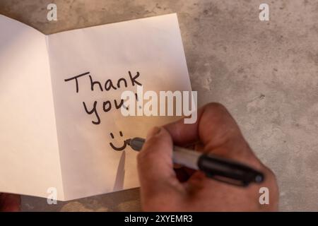Eine Person schreibt eine Dankesnotiz auf ein Blatt Papier. Die Notiz befindet sich auf einem Holztisch und die Person schreibt sie mit einem Marker. Szene ist positiv Stockfoto