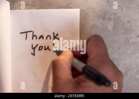 Eine Person schreibt eine Dankesnotiz auf ein Blatt Papier. Die Notiz befindet sich auf einem Holztisch und die Person schreibt sie mit einem Marker. Szene ist positiv Stockfoto
