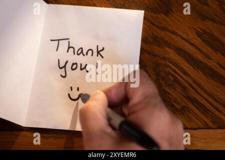 Eine Person schreibt eine Dankesnotiz auf ein Blatt Papier. Die Notiz befindet sich auf einem Holztisch und die Person schreibt sie mit einem Marker. Szene ist positiv Stockfoto