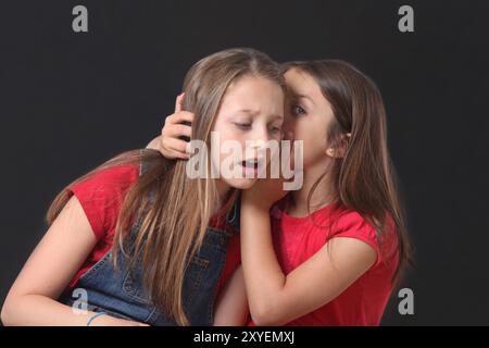 Zwei niedliche, stilvolle Mädchen, die über schwarzem Hintergrund tratschen. Das jüngere Mädchen flüstert ins Ohr eines älteren Mädchens. Stockfoto