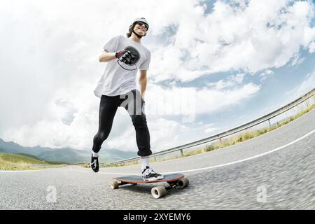 Ein Typ mit Helm und Sonnenbrille beschleunigt, indem er seinen Fuß auf seinem Longboard auf einer Landstraße auf Asphalt drückt und lächelt. Weitwinkel Stockfoto