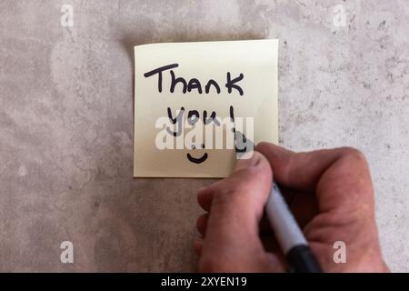 Eine Person schreibt eine Dankesnotiz auf ein Blatt Papier. Die Notiz befindet sich auf einem Holztisch und die Person schreibt sie mit einem Marker. Szene ist positiv Stockfoto
