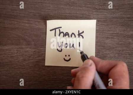 Eine Person schreibt eine Dankesnotiz auf ein Blatt Papier. Die Notiz befindet sich auf einem Holztisch und die Person schreibt sie mit einem Marker. Szene ist positiv Stockfoto